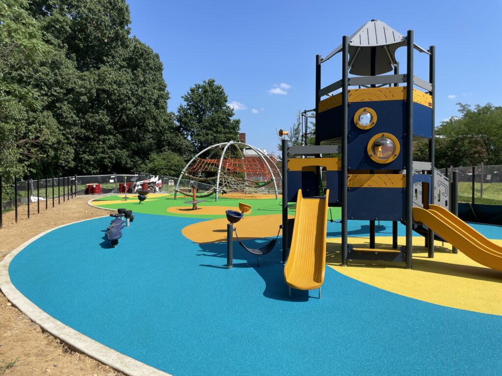 Custom Park Surfacing - Anne Beers second stage - view of playground structure with slide facing the viewer, large blue poured rubber surface nearest to the viewer, sunny day photograph.