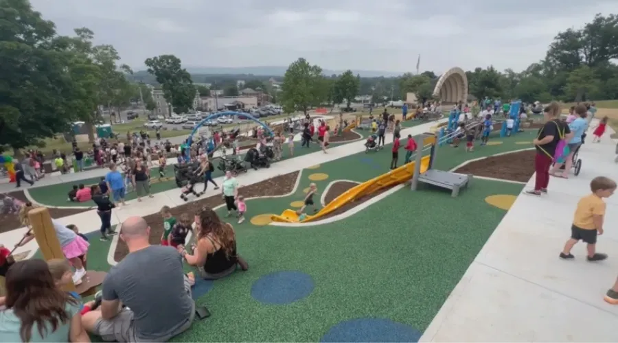Wide view of a crowded outdoor playground with winding slides, climbing structures, and families sitting on the turf as children play nearby.