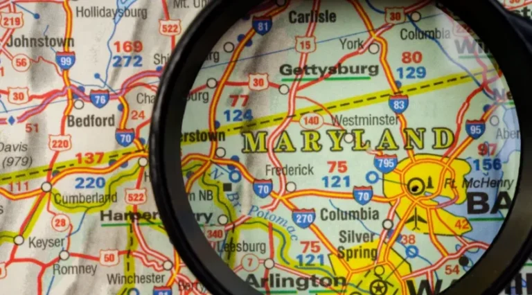 Close-up of a Maryland map with a black magnifying glass focusing on the Baltimore–Washington area, highlighting routes and cities like Baltimore and Columbia.